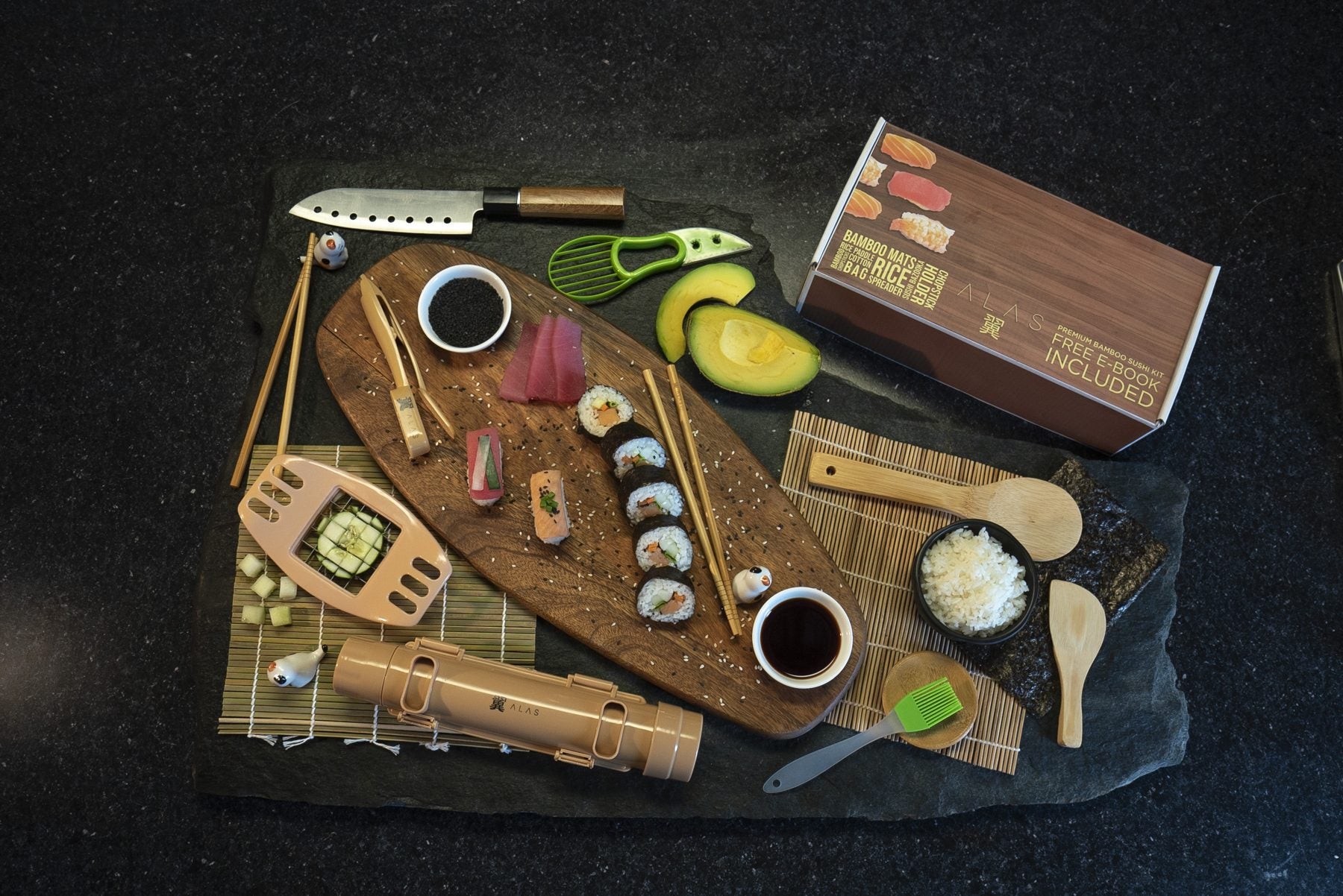 The full kit laid out on slate with a wooden board, sushi rolls, avocado and soy sauce