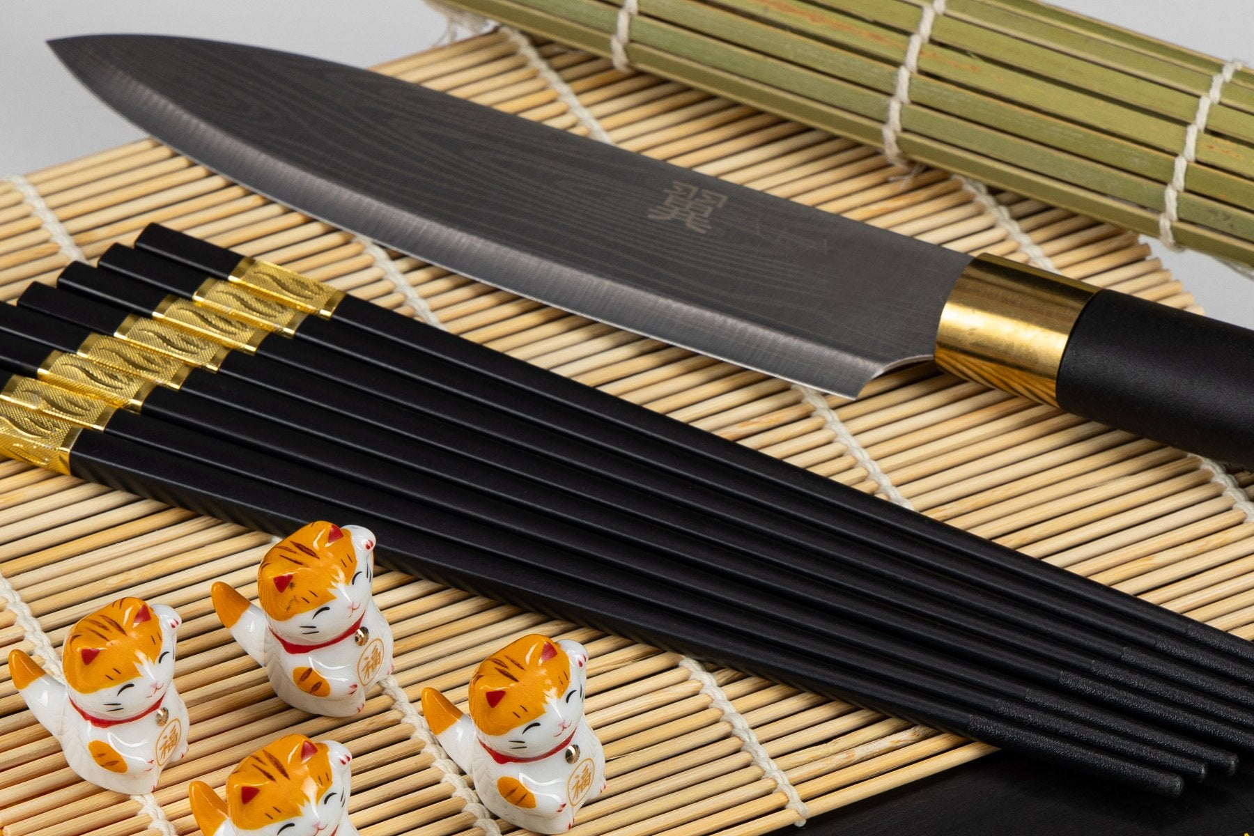 The black-handled sushi knife with black chopsticks and lucky cat rests on a bamboo mat